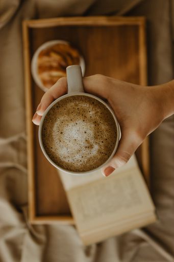 A man holding a cup of coffee on a blurred book and croissant on a wooden tray