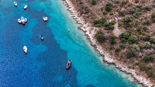 Aerial view of boats floating on clear blue waters next to a rocky coastline with lush greenery.