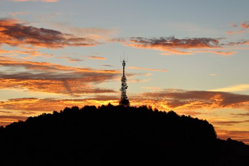 The silhouette of a transmission tower on a tree-covered hill under a colorful cloudy sky at sunset