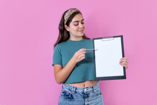 A woman pointing to a clipboard with a pen, posing in the pink background