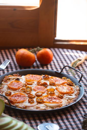 A freshly baked pizza topped with tomatoes and onions on a checkered tablecloth with fresh tomatoes in the background.
