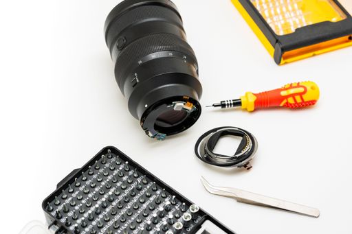 Broken camera lens with detached mount and precision tools on white background, showing professional photography equipment repair.