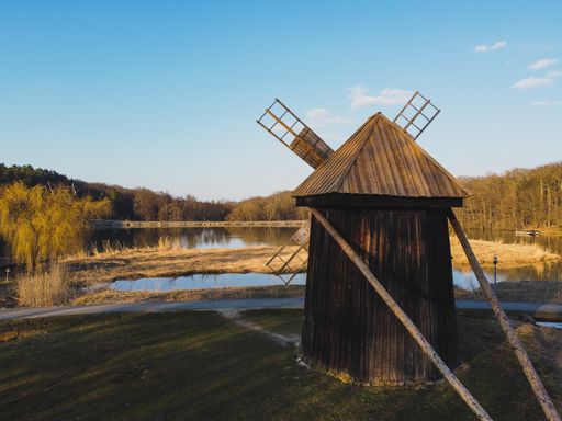An old wooden windmill by the reflective lake surrounded by trees under a blue clear sky in Romania