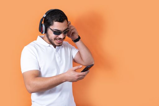 A young man with beard and sunglasses, listening to music with wireless headphones and smartphone; orange background with a copy space