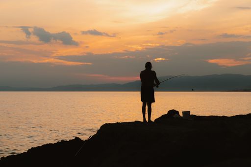 Silhouette of a person fishing at sunset by the sea, with an orange sky and distant mountains.