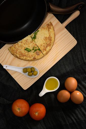 A vertical high angle closeup shot of two omelet pieces on a wooden cooking desk with olives, egg yolk, tomatoes, and fresh eggs on the sides