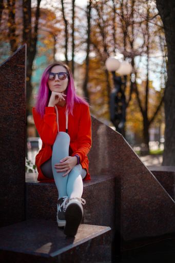 A vertical shot of a young girl with pink hair and sunglasses sitting on building stairs with a background of fall trees.