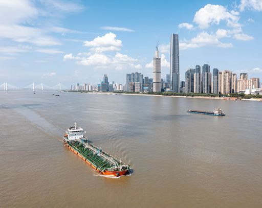 An oil transport ship navigates the Yangtze River with Wuhan's skyline in the background under a bright blue sky. Capturing urban development of China