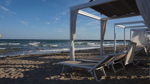 Beach cabanas and lounge chairs on a sandy shore with gentle waves and clear blue sky, perfect for a relaxing seaside day.