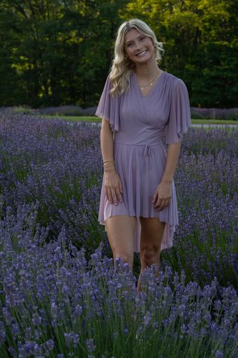 A young woman in a lavender dress stands joyfully in a lush lavender field on a sunny day.