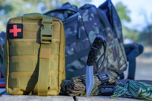 Tactical backpack with first aid kit, knife, and paracord lying on a wooden surface in nature