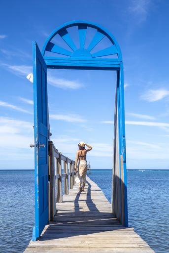 A beautiful vertical picture of a brunette female in a swimsuit behind of the blue door on a wooden bridge in Roatan Island