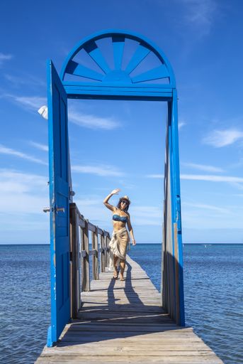 A beautiful vertical picture of a brunette female in a swimsuit behind of the blue door on a wooden bridge in Roatan Island