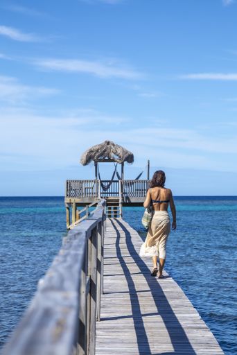 A beautiful vertical picture of a brunette female in a swimsuit walking on the wooden bridge against wooden hut on stilts in Roatan Island