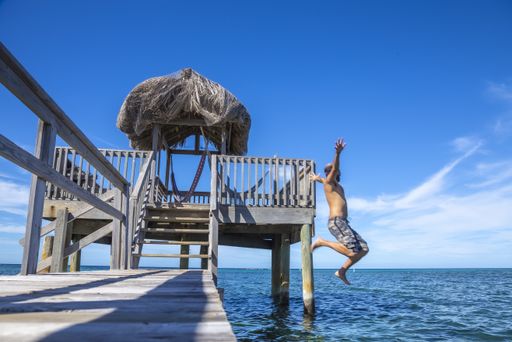 A beautiful picture of a brunette male in a swimsuit jumping in the water from the wooden bridge against wooden hut on stilts in Roatan Island