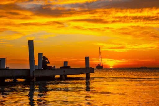 A beautiful picture of a silhouette sitting on the wooden bridge and taking a picture of a boat against a mesmerizing orange sunset