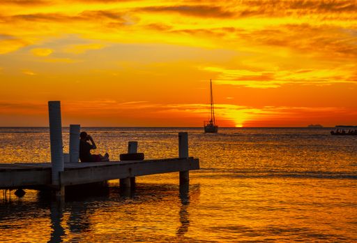 A beautiful picture of a silhouette sitting on the wooden bridge and taking a picture of a boat against a mesmerizing orange sunset