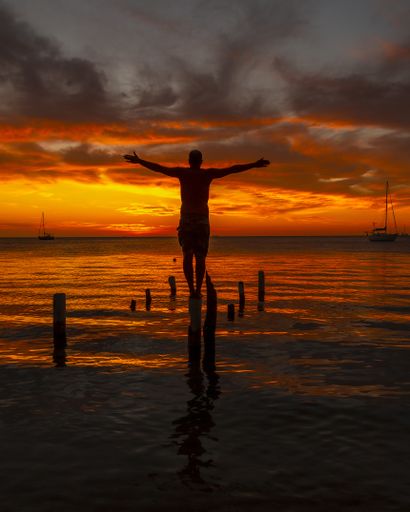 A beautiful vertical picture of a male silhouette standing on the wooden stilts on the water against an orange sunset in Roatan. Honduras