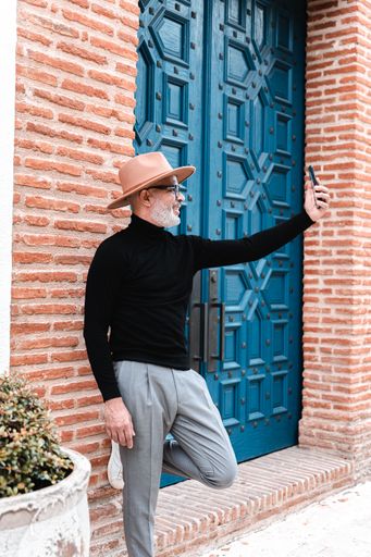 A stylish Hispanic male in eyeglasses and hat taking a selfie while leaning against a brick wall