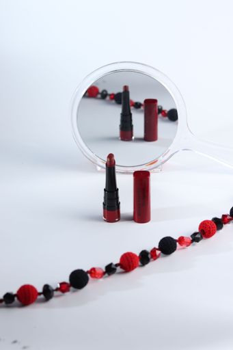 Red lipstick and beaded necklace reflected in a round mirror on a white background.