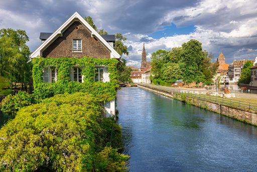 Scenic view of a vine-covered house by a river in Strasbourg, France, with a backdrop of historic architecture under a partly cloudy sky.