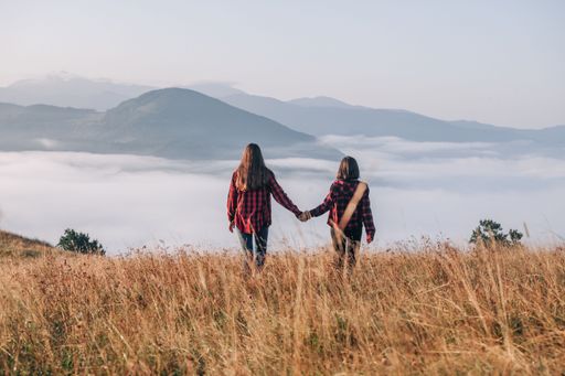 Two girls wearing red plaid shirts walk through a grassy mountain field under open sky.