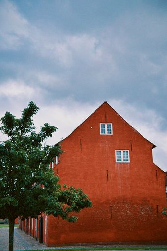 A vibrant red brick building with white windows stands against a cloudy sky, framed by a lush green tree.