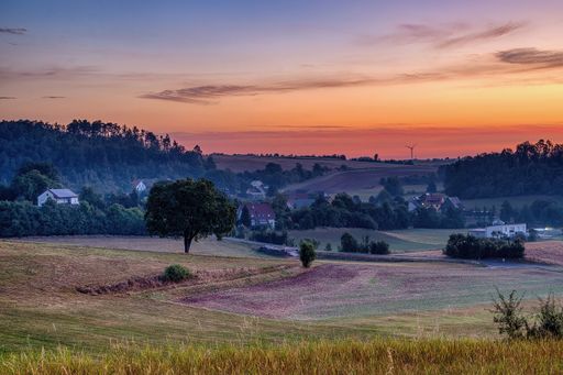 Scenic rural landscape at sunset with rolling hills, trees, and a wind turbine in the distance.