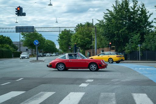 A classic red car at a city intersection with a yellow car and traffic lights under a cloudy sky.
