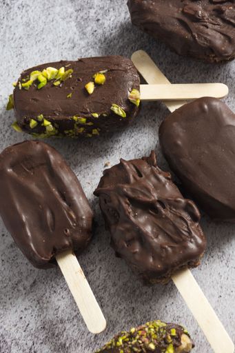 Chocolate-covered ice cream bars with pistachio topping on a textured surface, showcasing a delicious dessert arrangement.