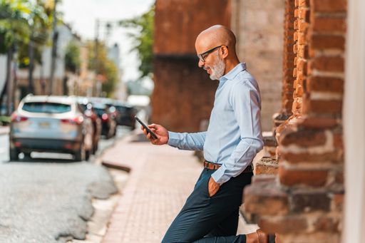 A bald Hispanic male using his smartphone while leaning against a brick wall