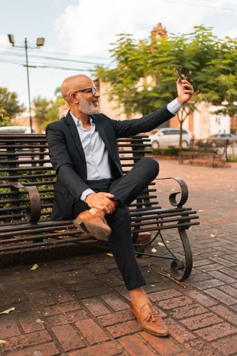 A handsome Hispanic male in an elegant suit talking a selfie while sitting on a bench