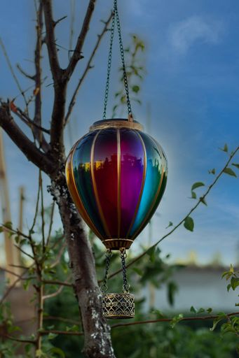 A colorful hot air balloon decoration hanging on a tree branch against a clear blue sky in a serene garden setting.
