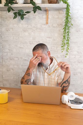 A middle-aged man with tattoos, holding glasses, looks stressed at a laptop in a modern home office setting.