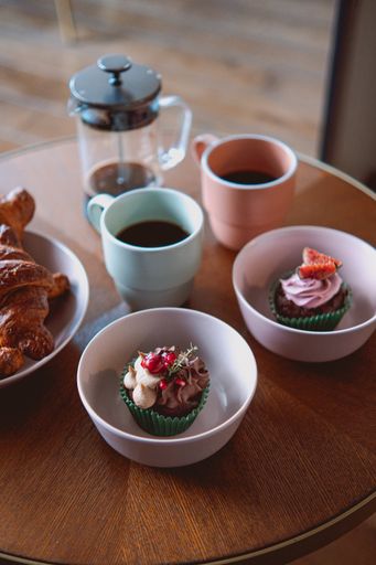 A vertical shot of cups of coffee with cupcakes and croissant bread in white bowls served in a cafe
