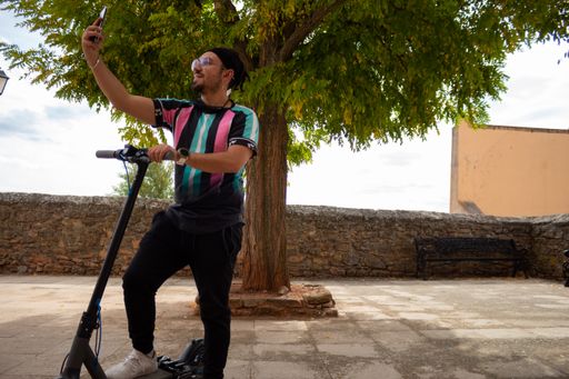 A young Caucasian male with glasses and braided hair taking a selfie with his electric scooter
