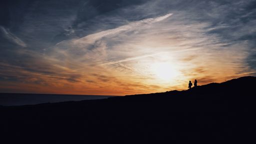 A wide shot of silhouettes of two people walking on a hill with the sun shining in the background