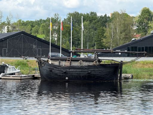 A dark, historic wooden ship is docked on a calm body of water, with flags and buildings visible in the background under a cloudy sky.