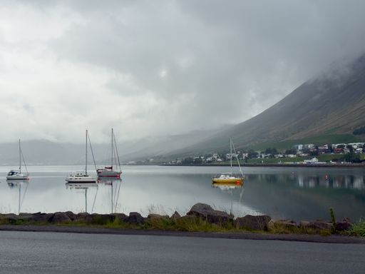 Serene coastal scene with sailboats on calm waters under a cloudy sky, mountains and a small town in the background.