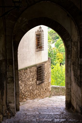 A scenic view through an ancient stone archway in a historic European village, revealing lush greenery and rustic architecture.