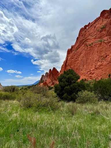 Dramatic red sandstone cliffs tower over grassy plains and scattered shrubs beneath a vivid blue sky and textured clouds.