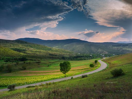 Scenic view of a winding road through lush green hills under a dramatic cloudy sky at sunset.