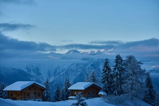 Scenic view of snow-covered cabins and pine trees against the backdrop of a majestic mountain range under a clear blue sky.
