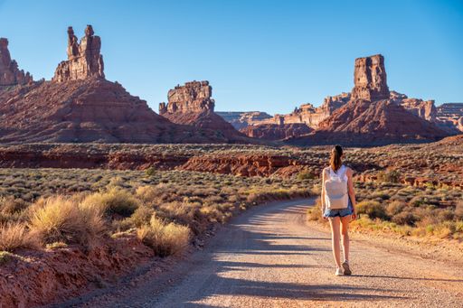 A young Caucasian woman walks down a dusty dirt road in the vast open desert, enjoying the scenery around her in Valley of the Gods.