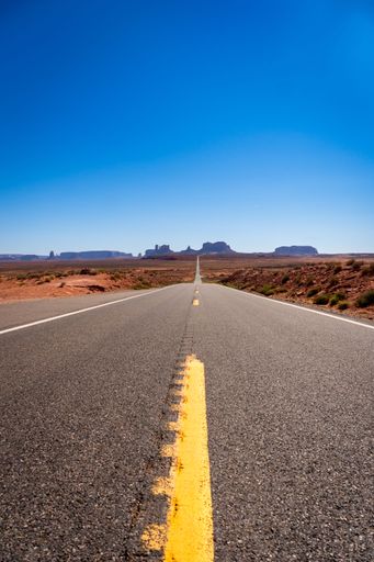A vertical shot of a long and straight road leads to distant mesas under a clear blue sky in Monument Valley, Arizona.