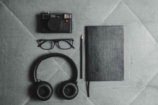 Flat lay of a camera, glasses, headphones, pencil, and notebook on a textured grey surface.