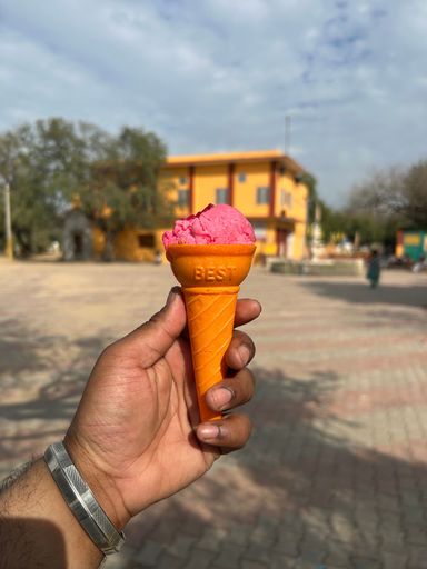 A close-up of a hand holding a pink ice cream cone outdoors with a building in the background under a cloudy sky.