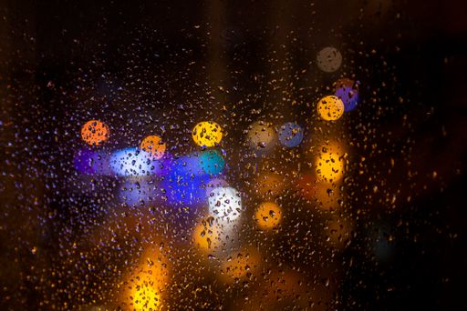 A closeup of a window covered in raindrops with a dark blurry background and bokeh lights