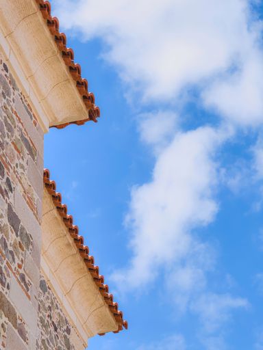 Close-up of a stone building corner with terracotta roof tiles against a bright blue sky with clouds.