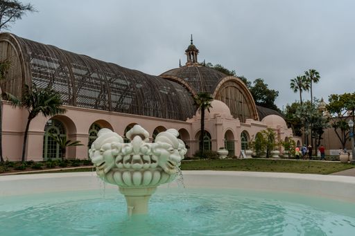 A beautiful ornate fountain is situated in front of a large, domed building with arches and a glass roof.